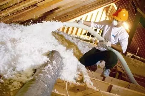 Technician wearing yellow hard hat, installing blown-in insulation in an attic.